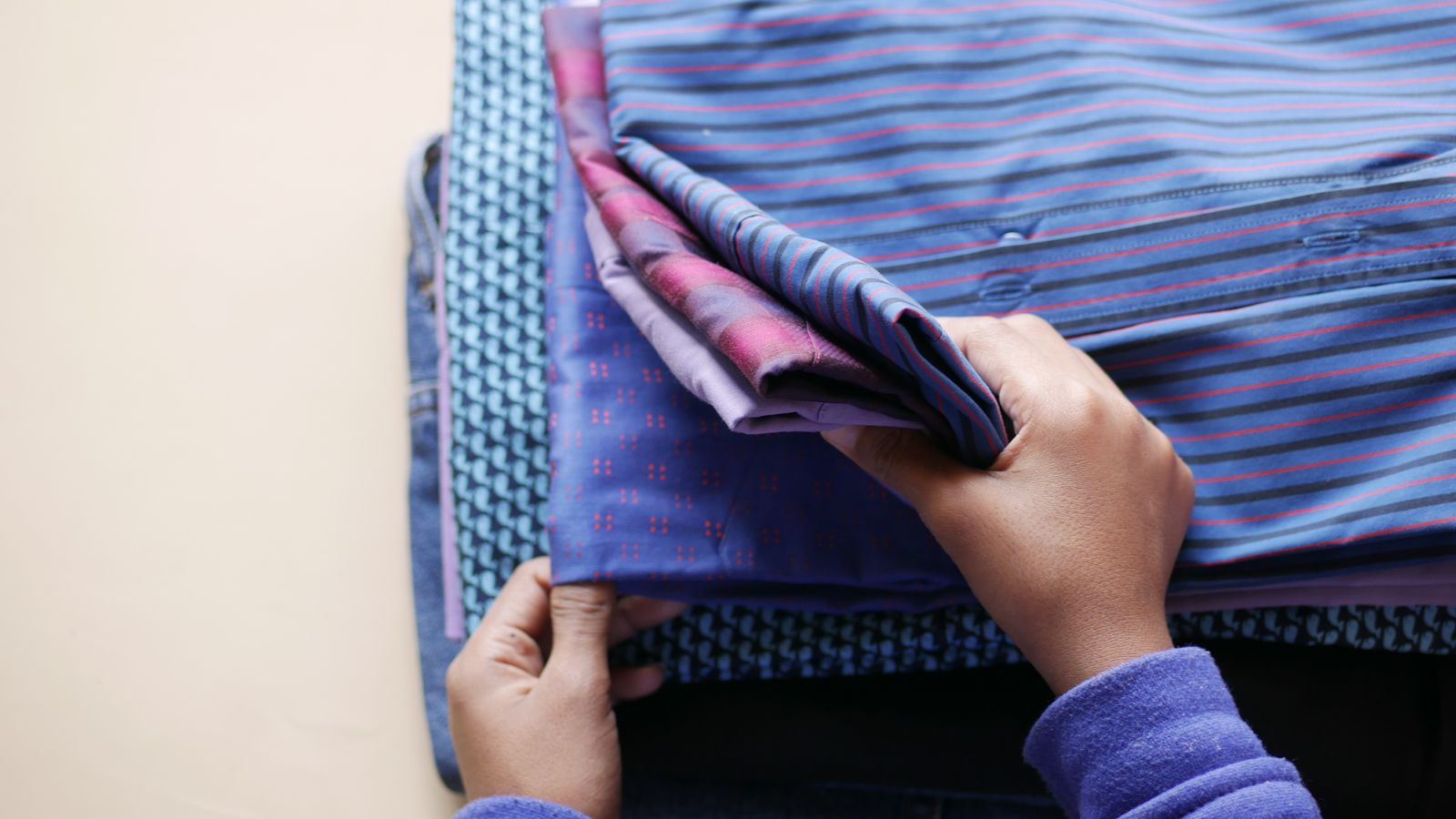 Mains pliant une pile de chemises colorées à motifs, disposées de manière soignée en vue aérienne.